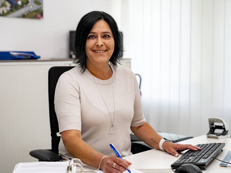 Christine Costa sitzt an einem Schreibtisch im Büro und lächelt in die Kamera