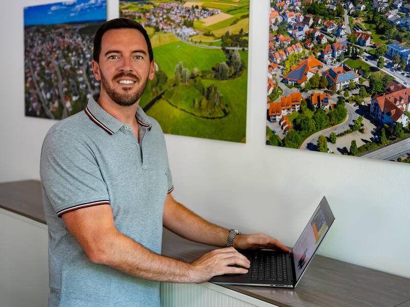 Florian Götz steht mit einem Laptop in einem Büro und lächelt in die Kamerea