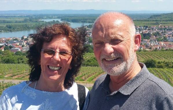 Ein lächelndes Paar mittleren Alters posiert für ein Foto vor einer hügeligen Landschaft mit grünen Weinbergen und einem blauen See im Hintergrund.