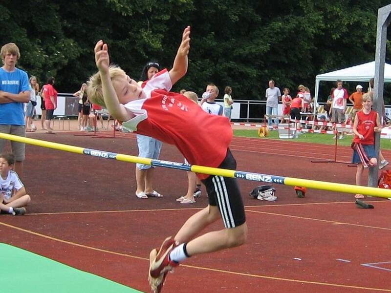 Ein Athlet in rotem Shirt und schwarzen Shorts beim Hochsprung über eine Latte, umgeben von Zuschauern im Hintergrund auf einem Sportplatz.