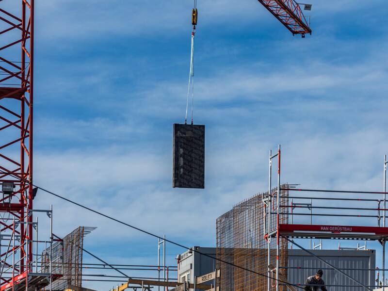 Ein Haus im Rohbau, das eingerüstet ist. Im linken Bildbereich befindet sich ein roten Kran,  welcher gerade Material an die Baustelle befördert.