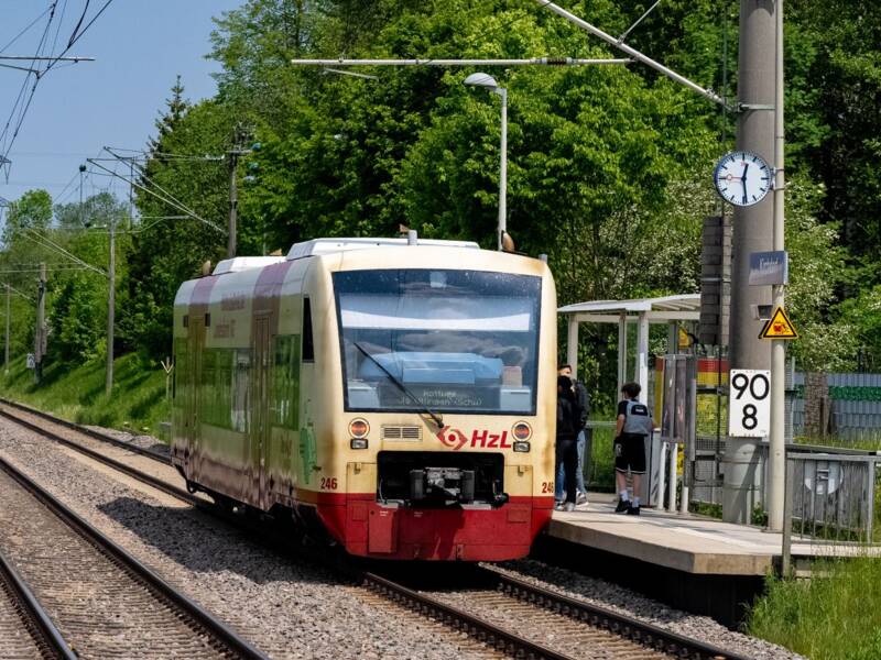 Ein roter und weißer Zug nähert sich auf einem Gleis, während im Hintergrund weitere Schienen und ein kleiner Ort sichtbar sind.