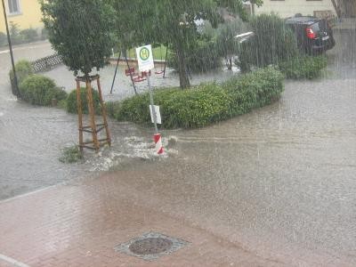 Auf einem gepflasterten Platz ist ein Bushaltestellenschild sowie Büsche und Bäume. Die Fläche ist überflutet und es regnet.