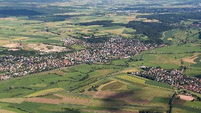 Luftbild der Gemeinde. Viele grüne und brauen Felder. In der Mitte sind Häuser sowie rechts am Bildrand. Hinten im Bild sind Wälder.